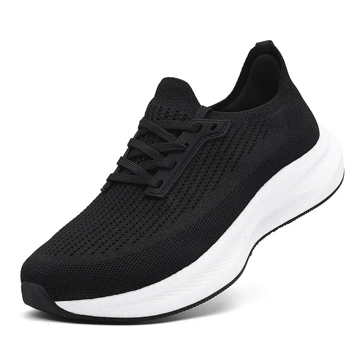 A black wide-fit athletic trainer with a breathable mesh upper, white EVA midsole, and black rubber outsole, shown from a three-quarter side angle against a studio background.