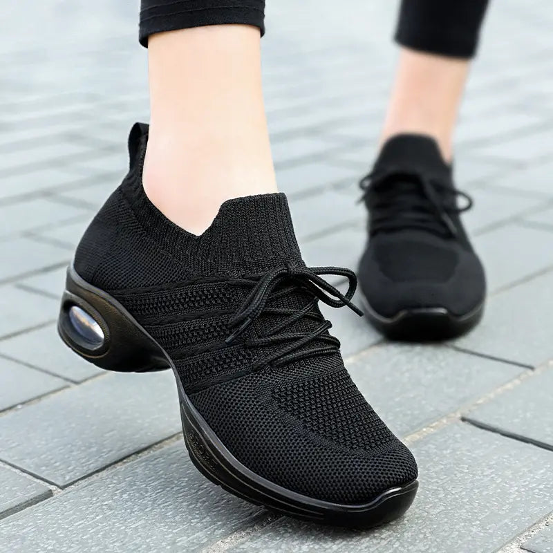 Women's black knitted athletic shoes with a clear air cushion sole and lace-up closure, shown from the side profile on a grey tiled surface.