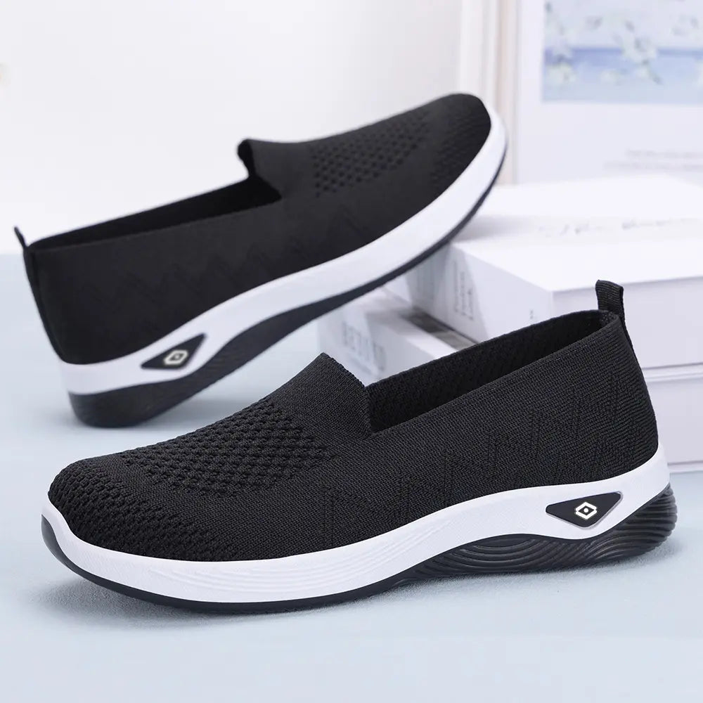 Black slip-on knit trainers for women, shown in profile view with white sculpted soles and a subtle debossed triangular logo on the heel.