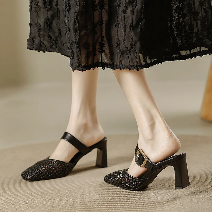 Model wearing black woven square toe block heel mules with an adjustable gold buckle strap, paired with a textured black dress on a neutral rug.