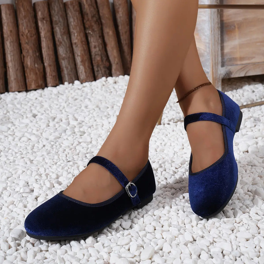 Deep sapphire blue velvet Mary Jane ballet flats with a delicate silver buckle strap, shown worn by a model on a decorative white pebble ground with a rustic wooden log backdrop.