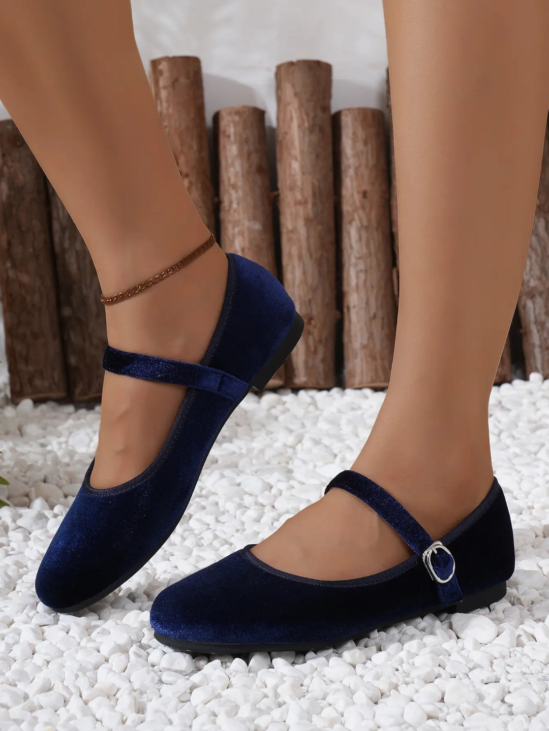 Blue velvet Mary Jane ballet flats with a silver buckle strap, worn on feet against a backdrop of white pebbles and rustic logs.
