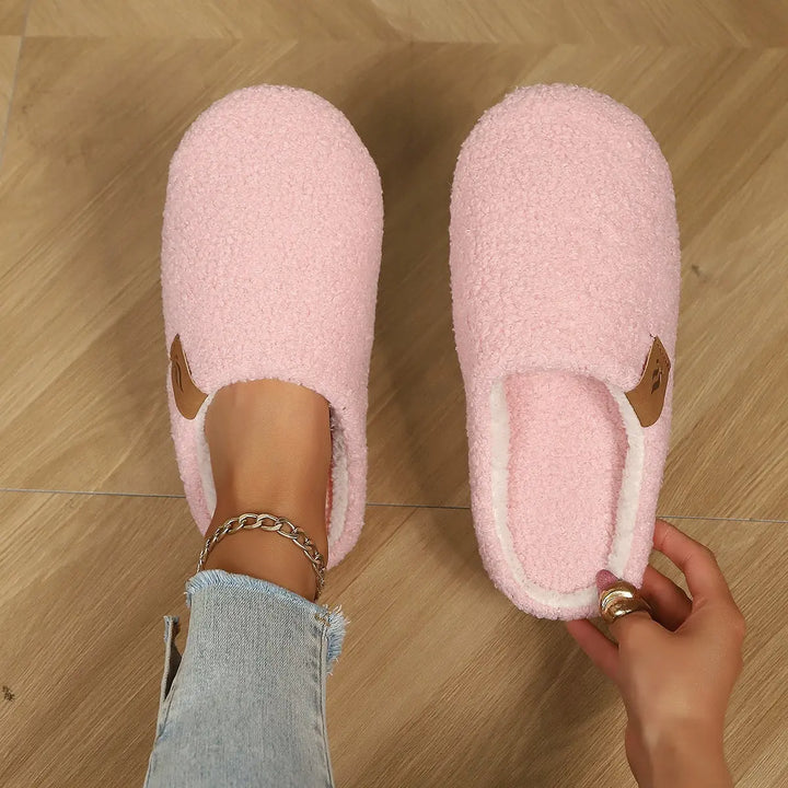 A pair of blush pink plush clog slippers worn by a person with a silver anklet and jeans, showcasing the soft texture and white lining, with a hand adjusting one slipper on a wooden floor.