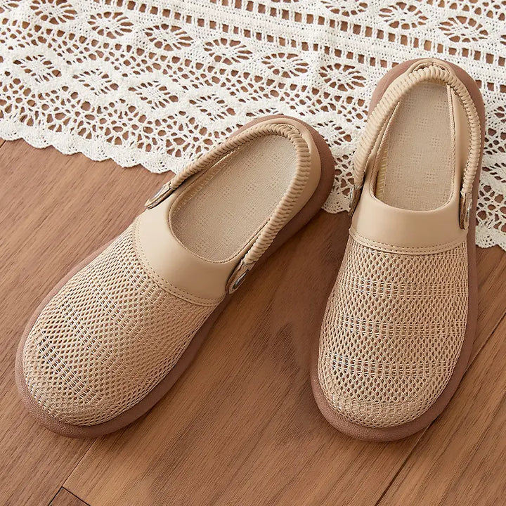 Pair of beige breathable mesh slip-on mules with synthetic trim and an elasticated heel strap, displayed on a wooden surface with a decorative lace overlay.