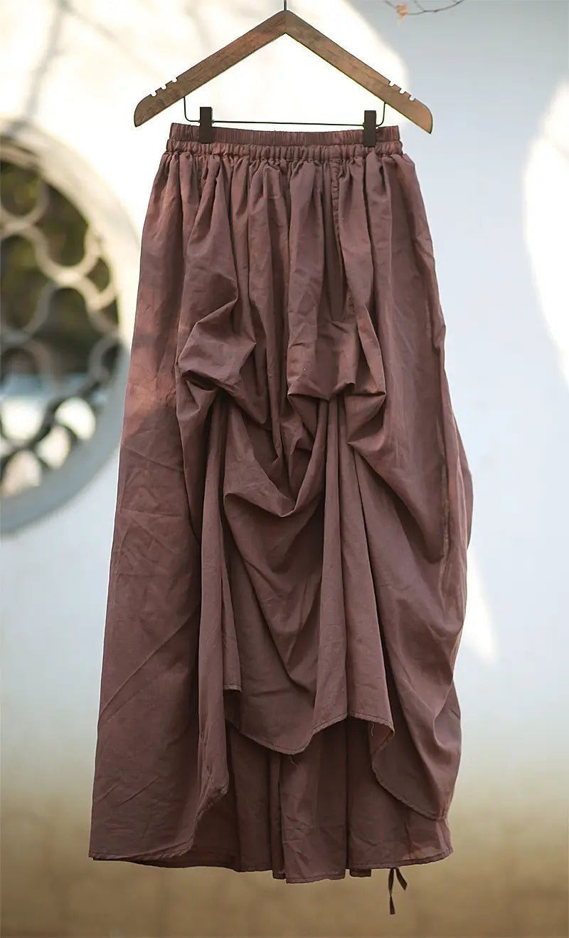Brown asymmetrical cotton maxi skirt with a draped silhouette and adjustable drawstring hem, hanging on a wooden hanger.