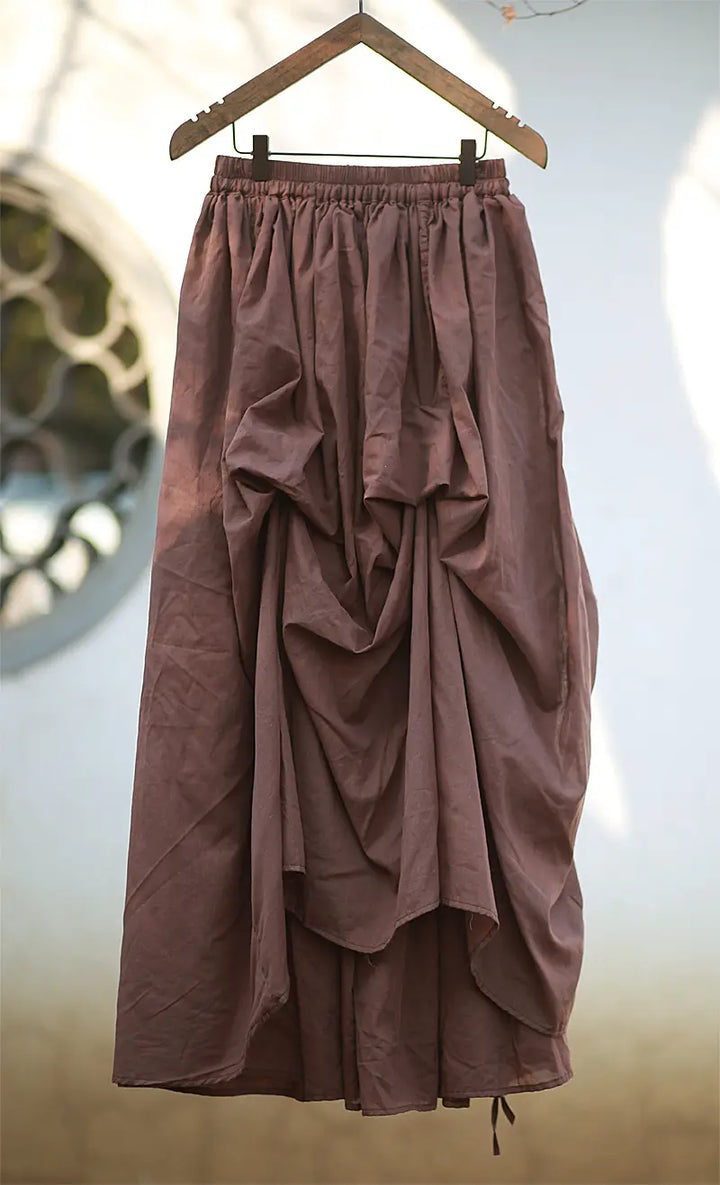 Brown asymmetrical cotton maxi skirt with a draped silhouette and adjustable drawstring hem, hanging on a wooden hanger.