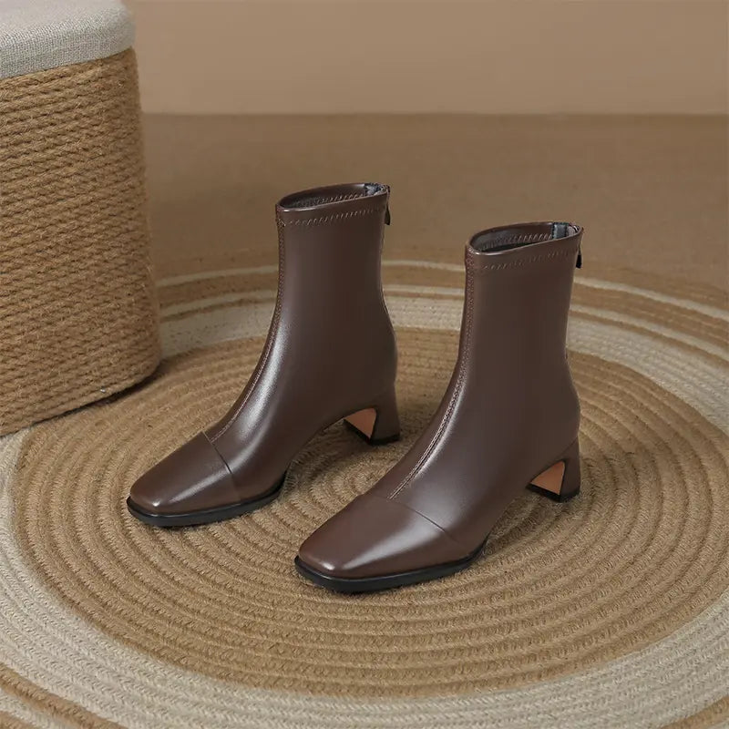 Brown faux leather square toe ankle boots with a block heel and interior side zipper, presented on a woven rug with a textured stool in the background.