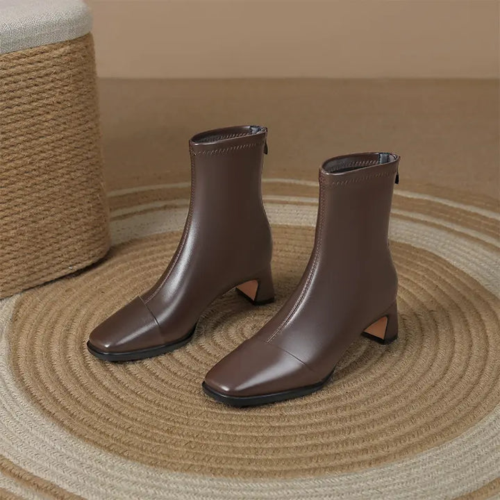 Brown faux leather square toe ankle boots with a block heel and interior side zipper, presented on a woven rug with a textured stool in the background.