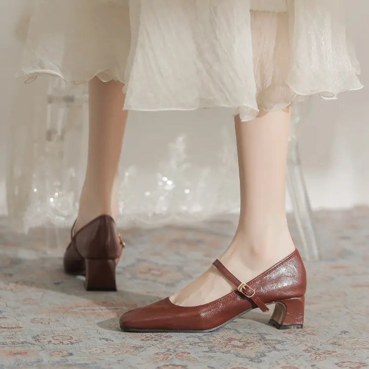 Chic brown Mary Jane block heel shoes with a square toe and single gold buckle strap, elegantly worn by a model with a flowing white skirt.