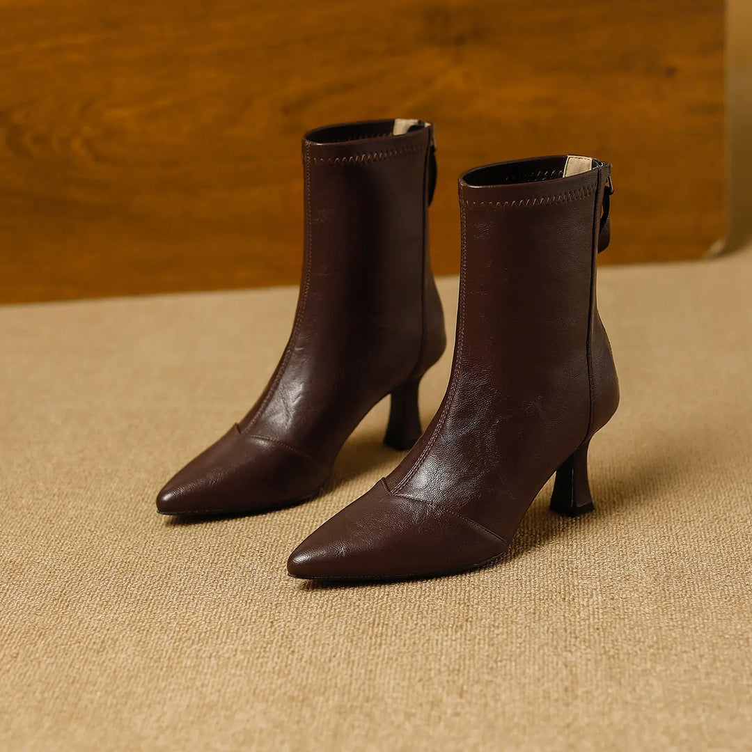 A pair of rich brown faux leather ankle boots with a sharply pointed toe and back zipper closure, showcased on a textured studio floor.