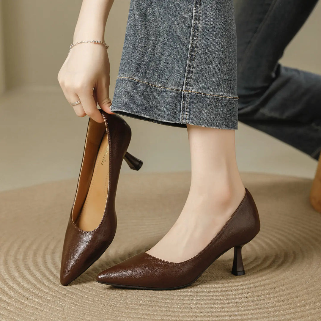 Brown faux leather pointed-toe pumps with a flared kitten heel, shown from a three-quarter angle as a hand holds one shoe and the other is worn.