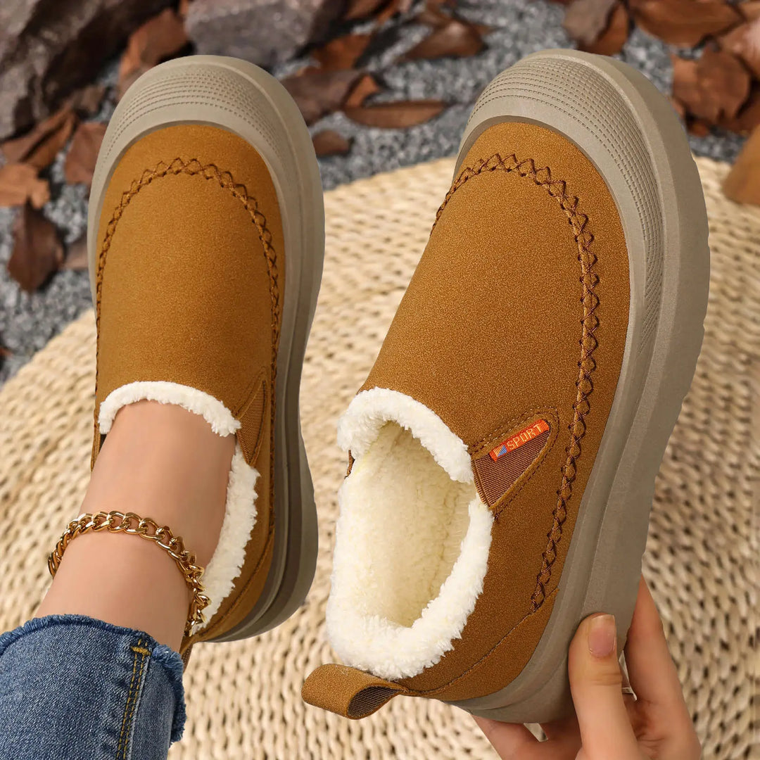Brown suede-like slip-on winter boots with plush shearling lining, decorative stitching, and a chunky grey platform sole, styled on feet with cuffed jeans.