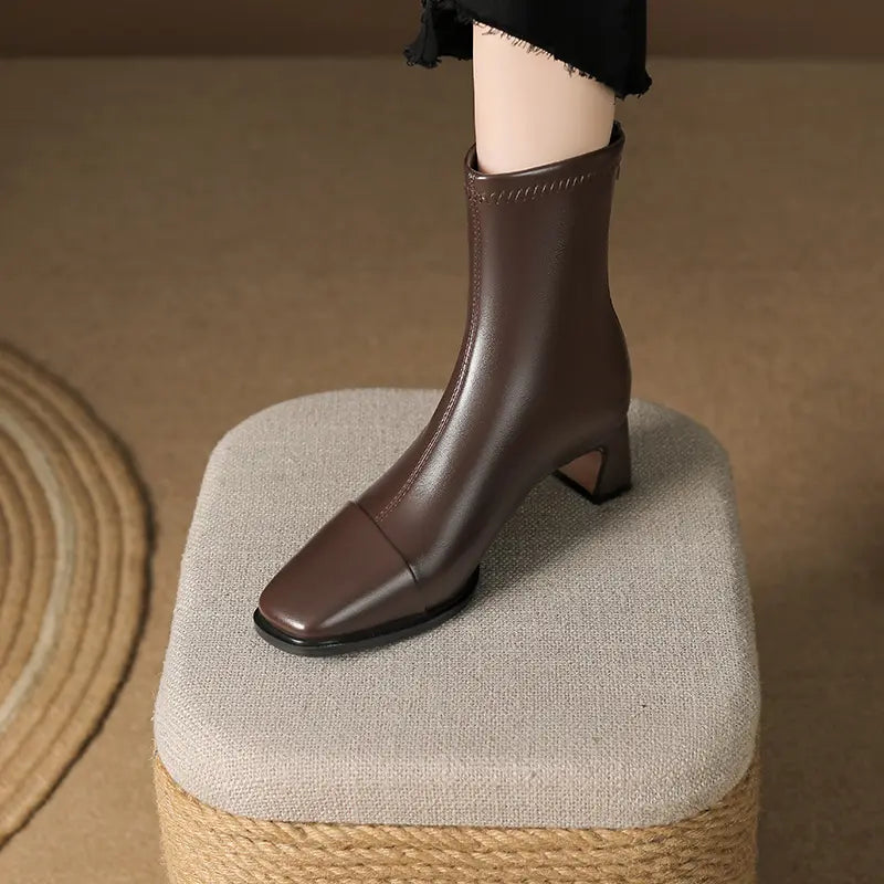 Rich brown faux leather ankle boot with a chic square toe and stable block heel, shown from a three-quarter side view on a decorative stool.