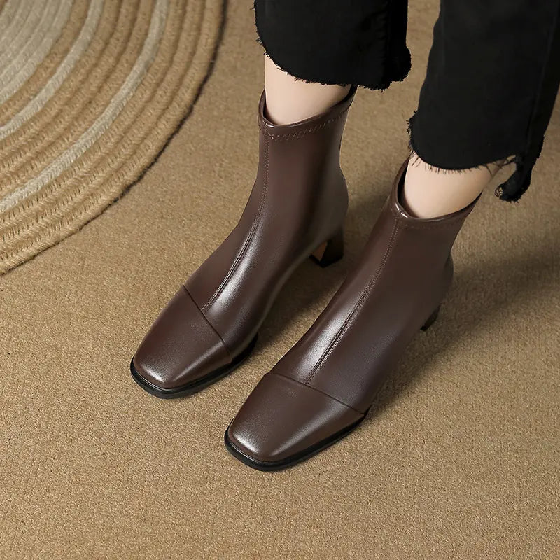 Brown faux leather square toe ankle boots with a block heel, shown on a person wearing black pants on a textured floor.