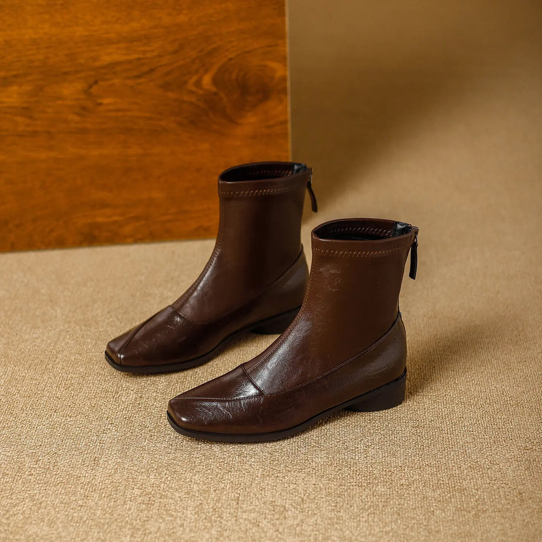 Brown leather-look ankle boots with a square toe, low block heel, and back zipper, presented in a studio setting with textured flooring and a wooden panel.