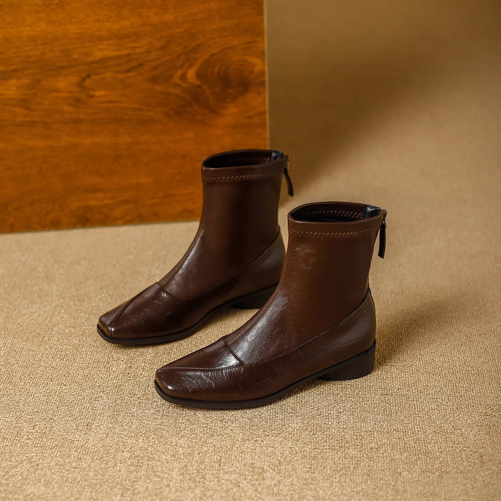 Brown leather-look ankle boots with a square toe, low block heel, and back zipper, presented in a studio setting with textured flooring and a wooden panel.