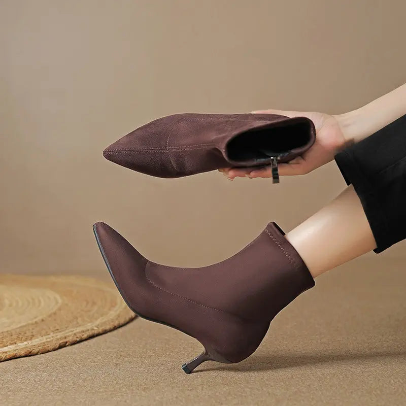 A pair of dark brown suede-like ankle boots with pointed toes and slender stiletto heels, one worn on a foot and the other held by a hand. The boots feature a stretch sock-like shaft and an inner side zipper. Displayed on a textured background.