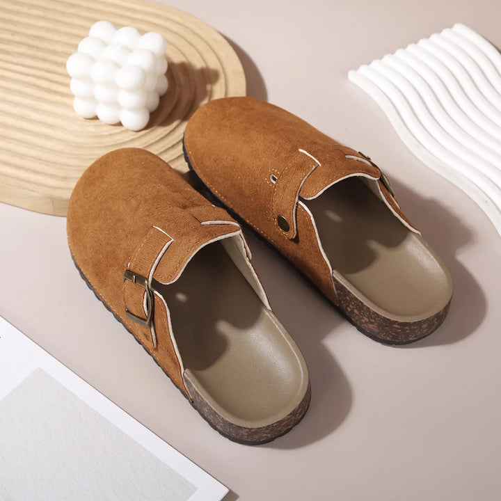 A pair of brown suede-like mules with adjustable brass buckles, featuring a cork-like midsole and a plush faux shearling lining, displayed from a three-quarter angle on a studio surface with decorative elements.
