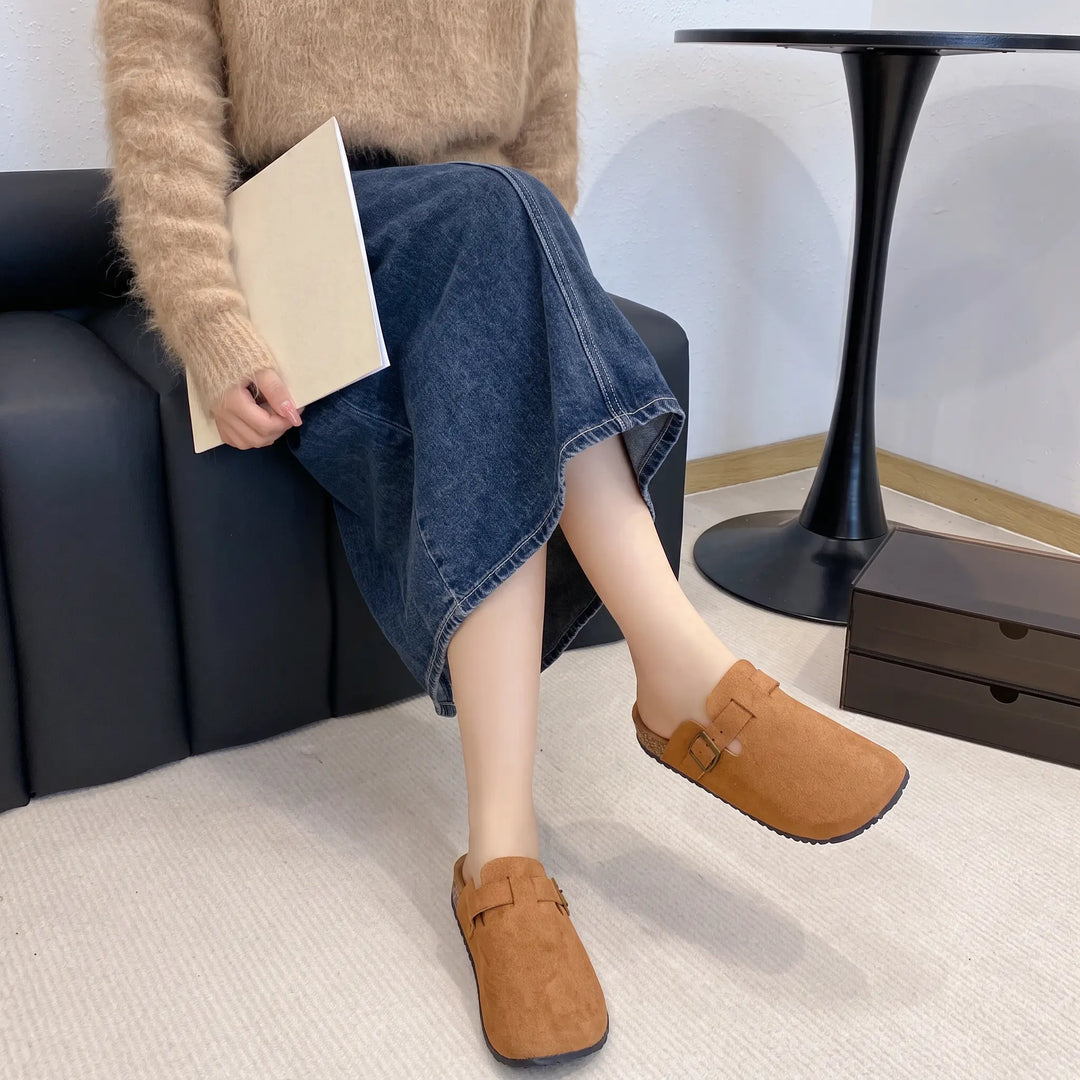 Woman wearing brown suede-like closed-toe mules with faux shearling lining and adjustable brass buckle straps, styled with a denim midi skirt and fuzzy sweater.