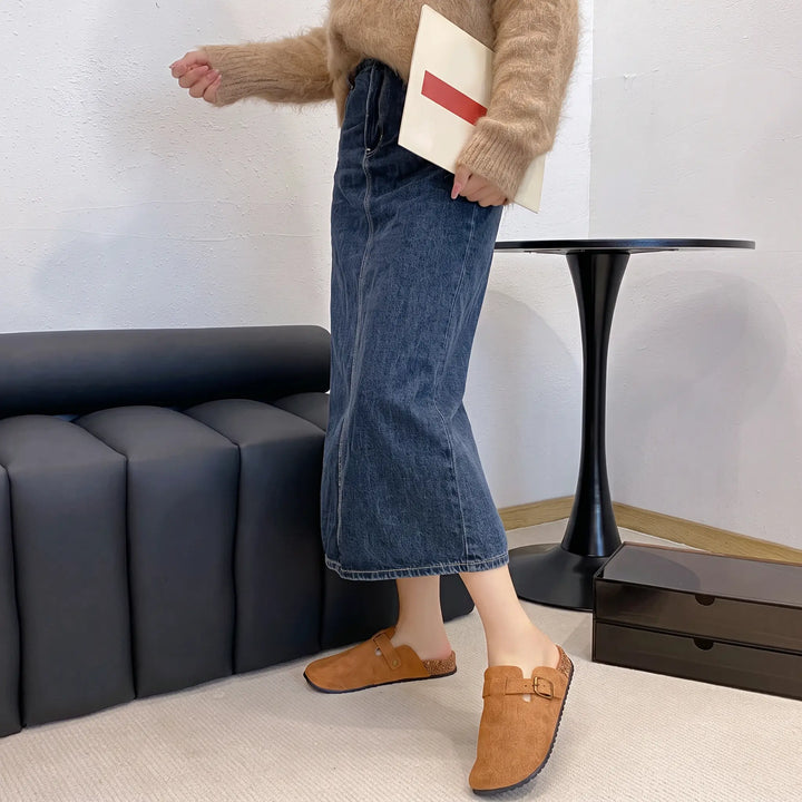A woman wearing brown suede-like mules with an adjustable buckle strap and faux shearling lining, styled with a denim midi skirt and a cozy sweater.