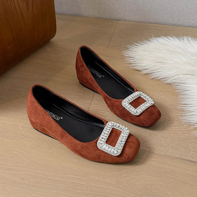 Brown suede slip-on wedge flats for women, featuring a prominent square buckle encrusted with clear faceted crystals on the vamp. These elegant shoes offer a rounded-square toe and a subtle wedge heel.