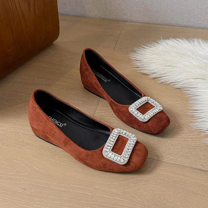 Brown suede slip-on wedge flats for women, featuring a prominent square buckle encrusted with clear faceted crystals on the vamp. These elegant shoes offer a rounded-square toe and a subtle wedge heel.