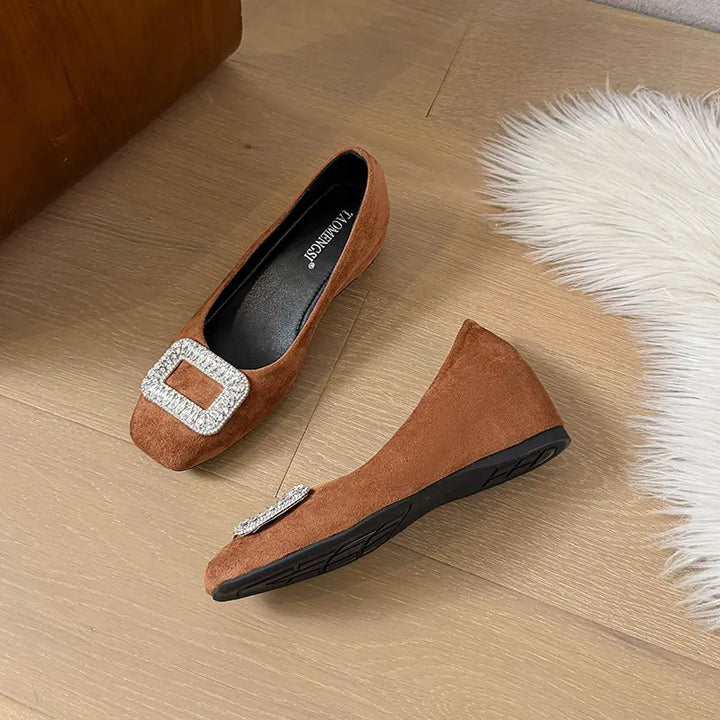 Brown suede slip-on wedge flats adorned with a large, rectangular crystal-embellished buckle on the vamp, showcasing a rounded-square toe and a subtle wedge heel.