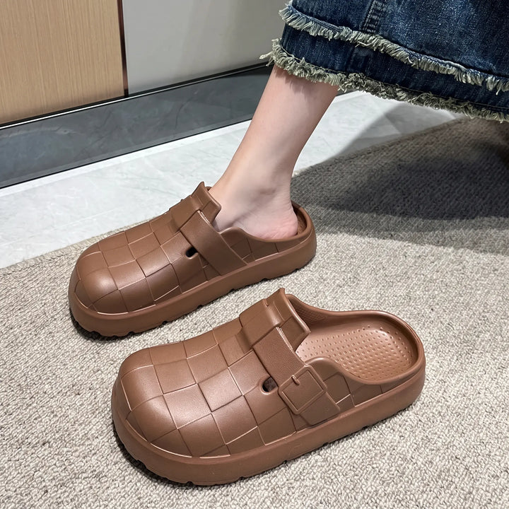A person wearing brown woven EVA clogs with an open back and thick platform sole, shown in a lifestyle context on a rug.