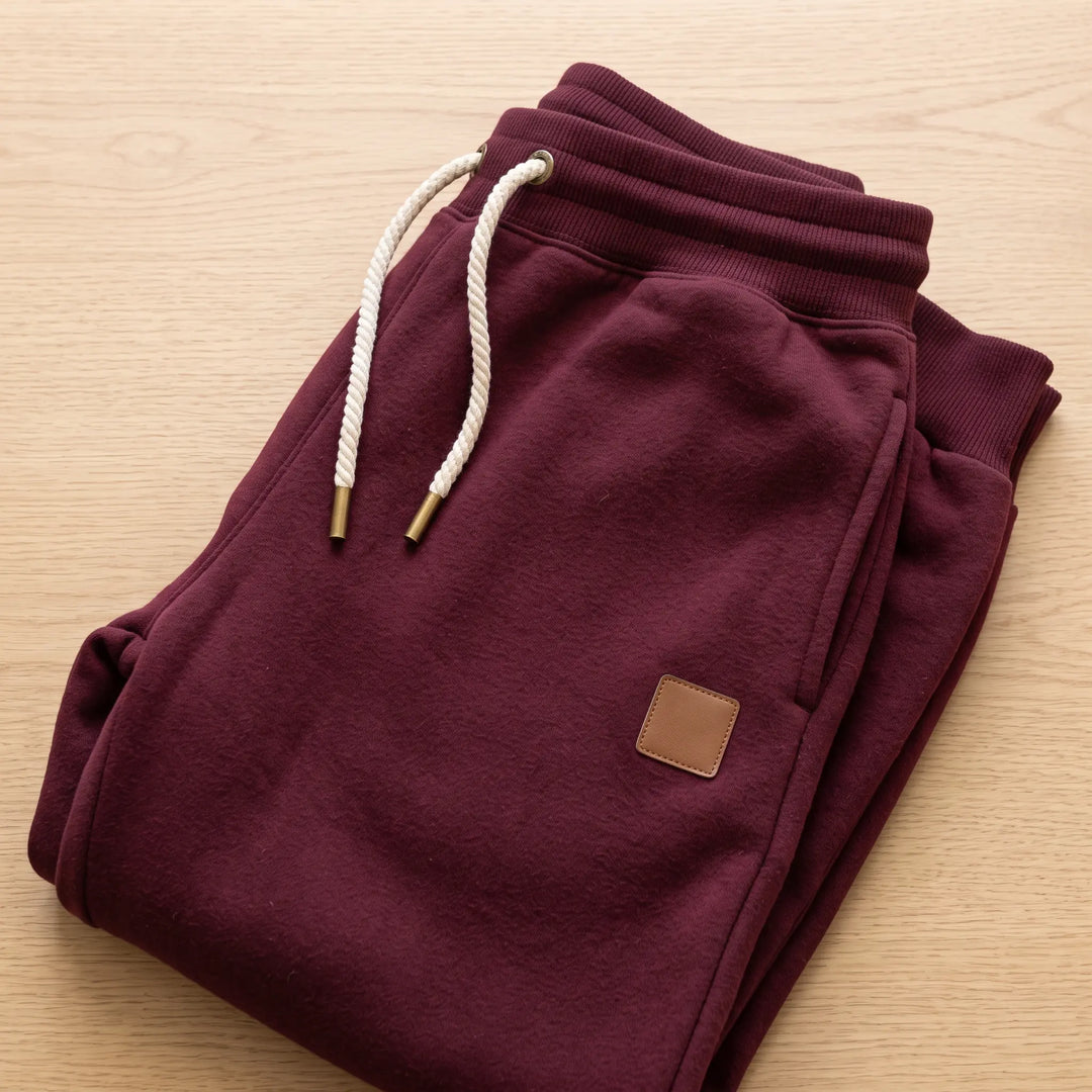 Burgundy tapered fit fleece-lined joggers with ribbed cuffs, drawstring waist, side pockets, and a subtle faux-leather logo patch on the thigh.