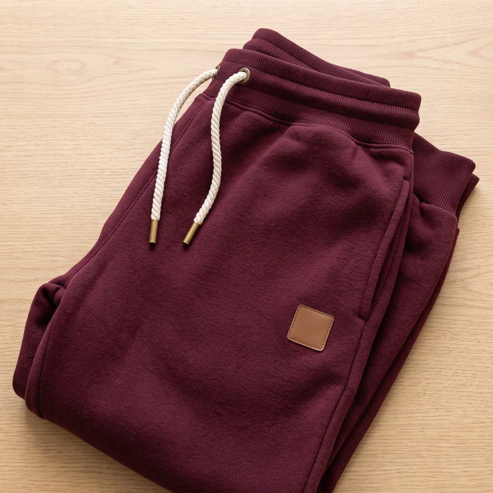 Burgundy tapered fit fleece-lined joggers with ribbed cuffs, drawstring waist, side pockets, and a subtle faux-leather logo patch on the thigh.