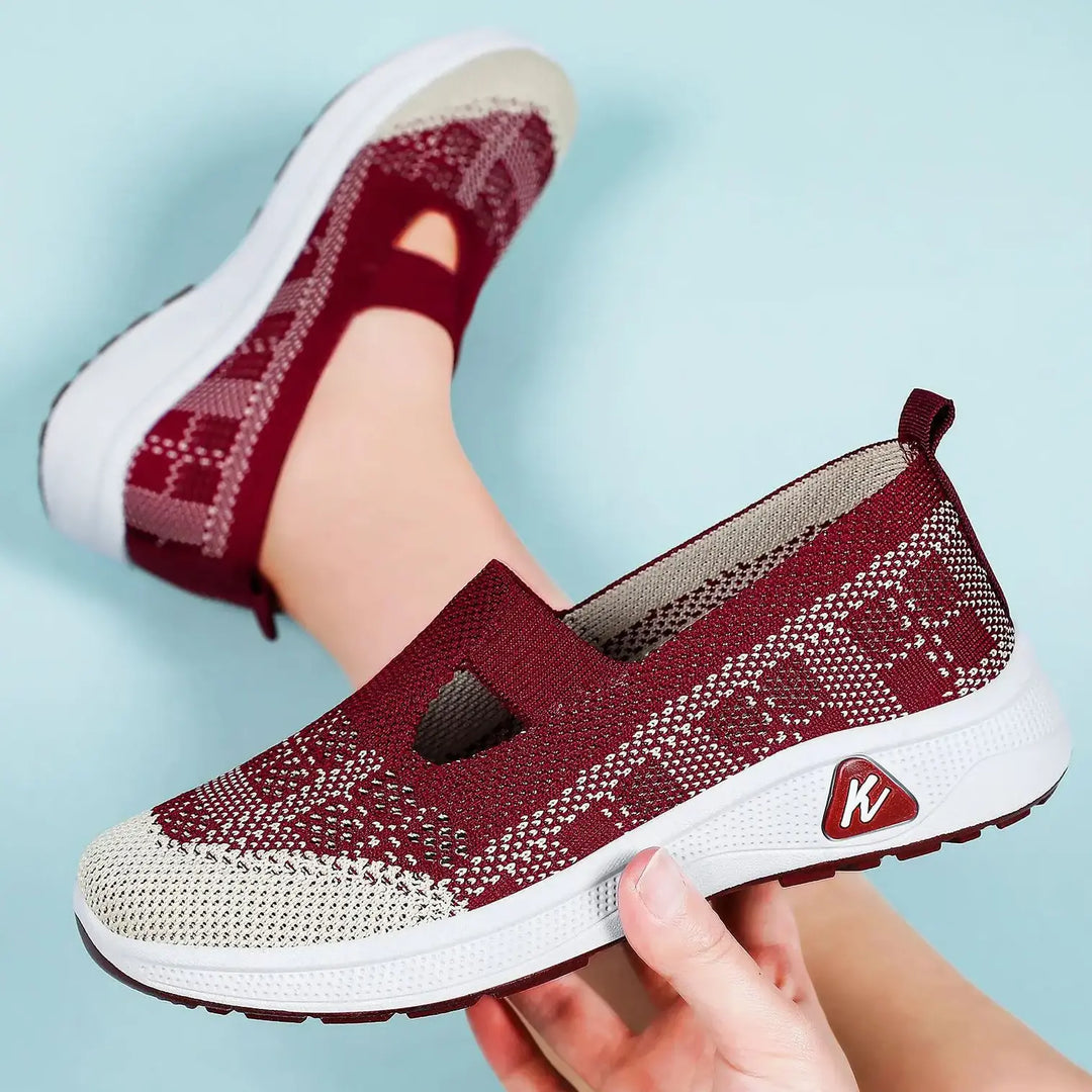 Three-quarter view of burgundy and beige geometric mesh slip-on sneakers shown on a foot and held by a hand, featuring a keyhole instep cutout and a white sole with pink accents.