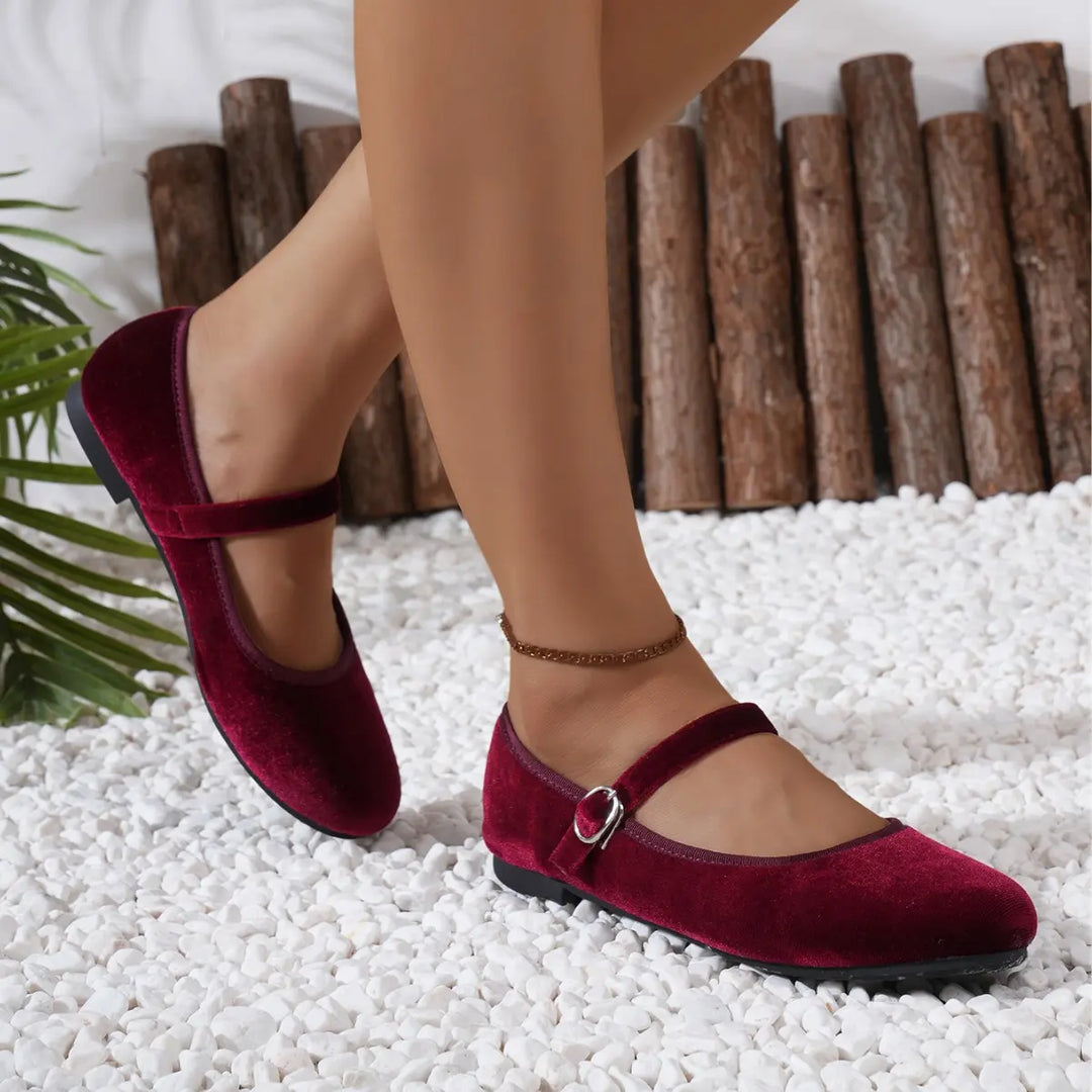 Burgundy velvet Mary Jane ballet flats with an adjustable buckle strap, worn on feet in a lifestyle setting with white pebbles and a wooden log backdrop.