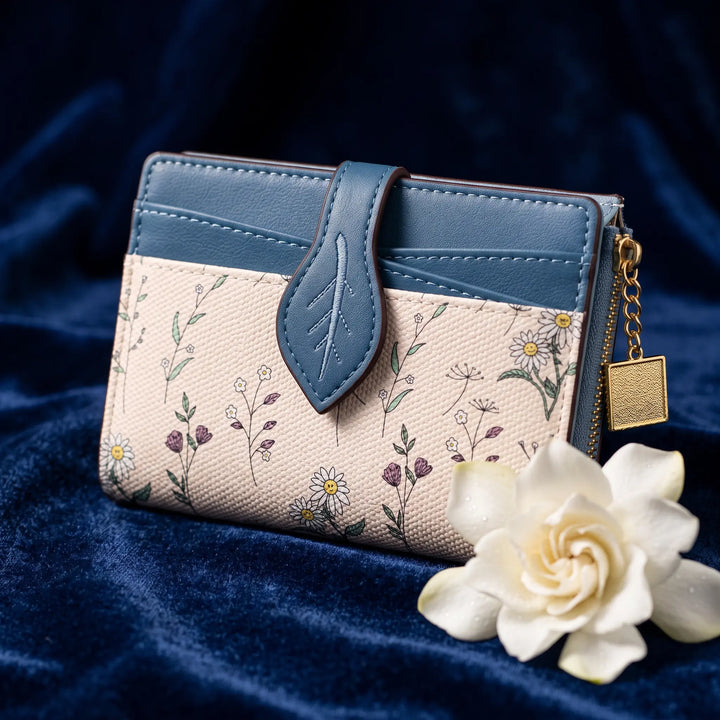 Compact bi-fold wallet featuring an off-white base with a delicate floral print, a contrasting blue leaf-shaped tab closure, and gold-tone zipper detail, presented with a white flower on a textured blue background.