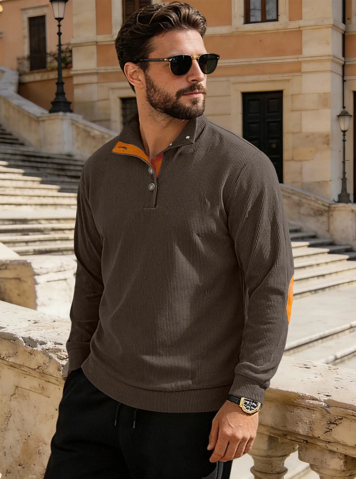 Man wearing a dark grey fine-ribbed corduroy Henley pullover with orange collar trim, tan suede-like elbow patches, and antique brass buttons, set in an outdoor European street scene.