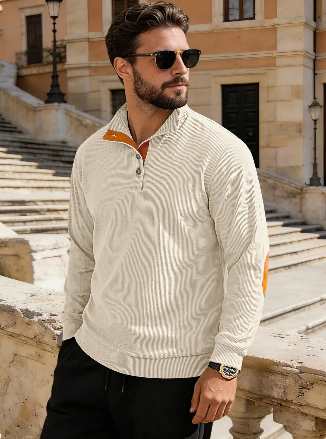 Man wearing a cream fine-ribbed corduroy Henley pullover with stand collar, button placket, and tan suede elbow patches, standing outdoors.