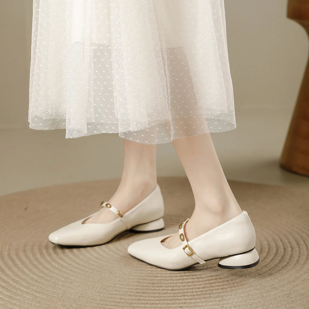 Cream square-toe Mary Jane shoes with a low block heel and gold stud buckle strap, shown on a foot against a textured rug.