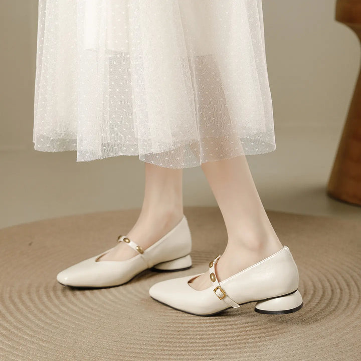 Cream square-toe Mary Jane shoes with a low block heel and gold stud buckle strap, shown on a foot against a textured rug.