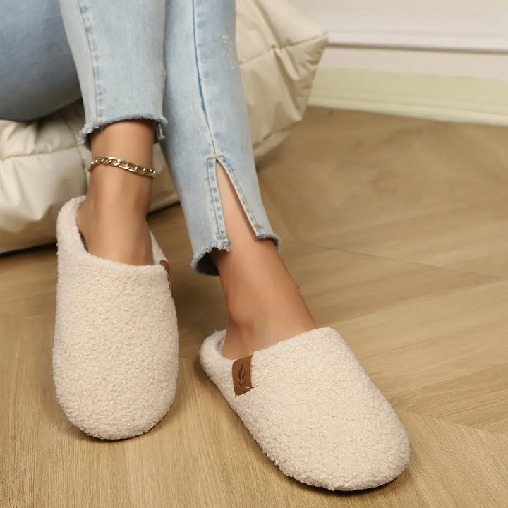 Cream beige plush clog slippers with white faux fur lining, worn by a person in light blue jeans and an ankle bracelet on a wooden floor.