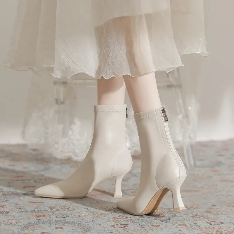 Creamy off-white pointed-toe stiletto ankle boots shown on legs wearing a flowing white dress, styled in a lifestyle setting on a patterned rug.