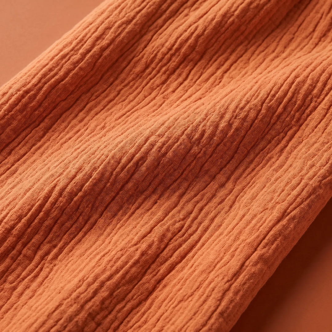 Close-up detail of warm orange crinkled cotton gauze fabric, showcasing its unique texture and breathable weave for a midi wrap dress.
