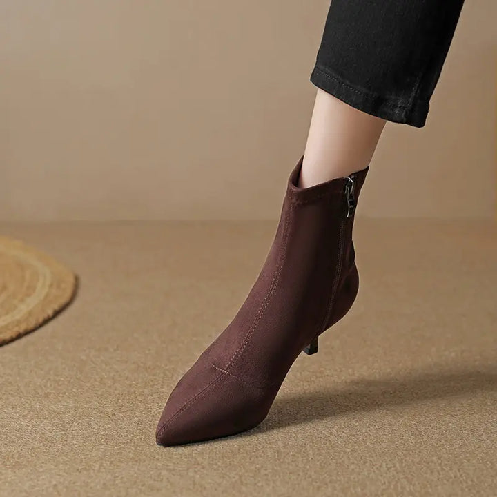A dark brown suede-like stiletto ankle boot with a pointed toe and side zipper is worn on a person&