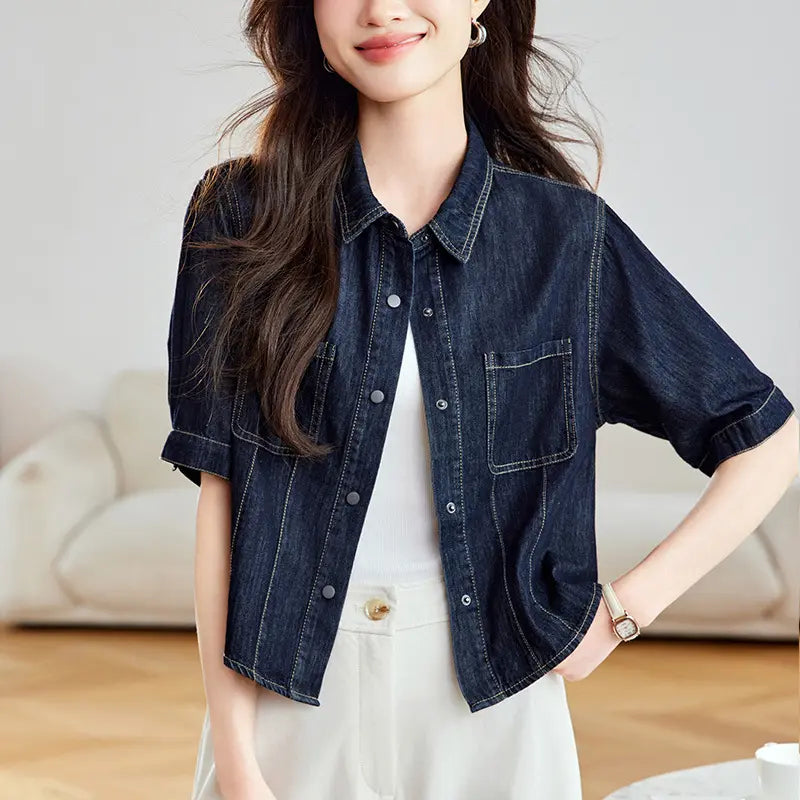 Model wearing a dark wash denim shirt jacket with short sleeves, contrast stitching, and silver snap buttons, paired with cream trousers.