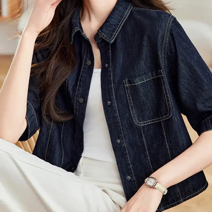 Model wearing a dark wash cropped denim shirt jacket with short sleeves, contrast stitching, and silver snap buttons, styled with a white tank top and cream trousers.