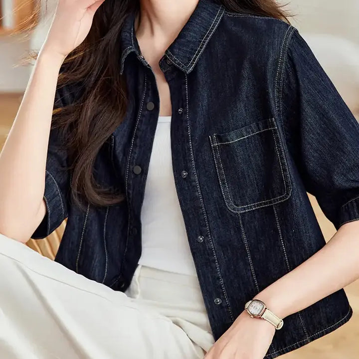 Model wearing a dark wash cropped denim shirt jacket with short sleeves, contrast stitching, and silver snap buttons, styled with a white tank top and cream trousers.