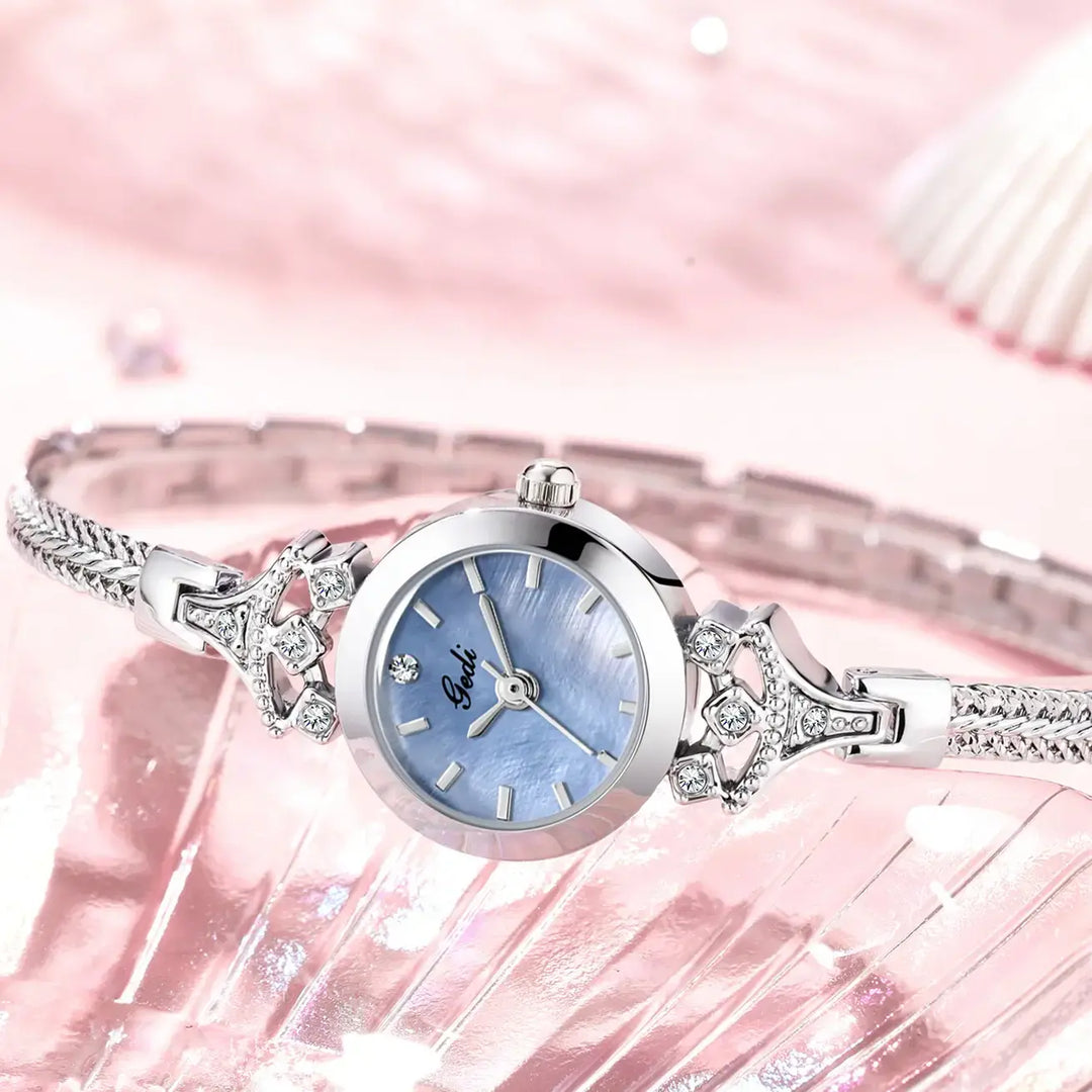 Elegant silver-tone bracelet watch with a delicate blue mother-of-pearl dial and crystal-embellished lugs, presented from a slightly elevated angle on a soft pink background.