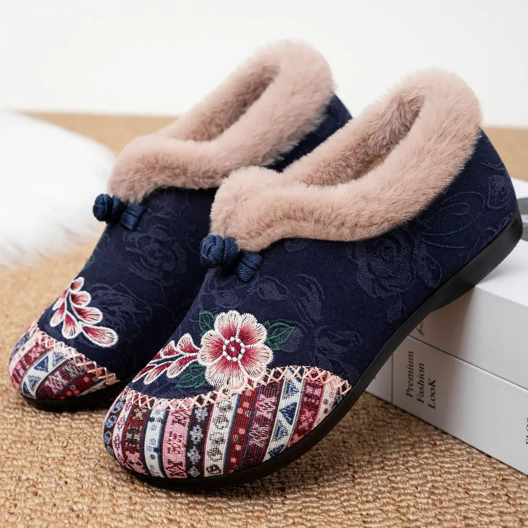 Pair of navy blue embroidered mules with colorful floral and geometric patterns, plush light brown faux-fur trim, and small dark grey bows.