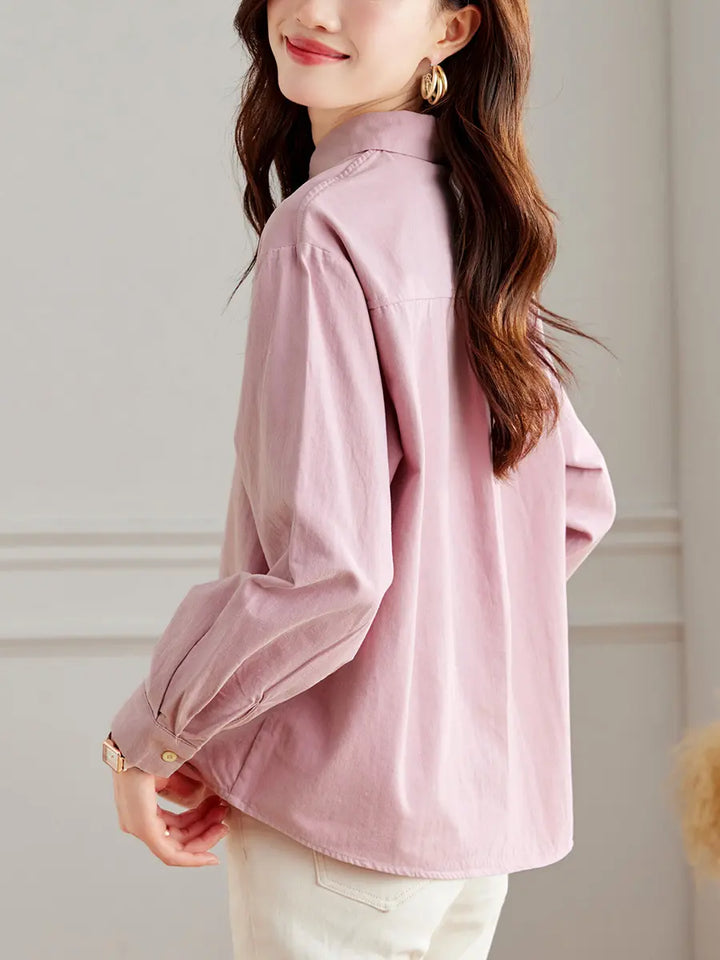 A woman in a muted pink embroidered cotton shirt, shown from a three-quarter back view, styled with light-colored trousers in a studio setting. Features include a classic collar and button-down front.