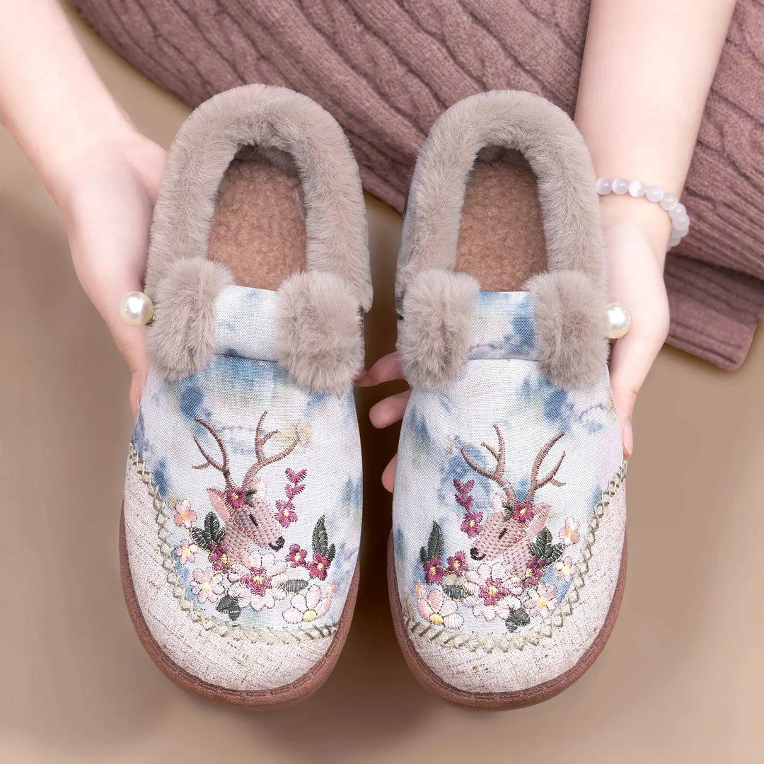 Embroidered plush slippers in soft pinkish-mauve with floral and deer motifs, featuring grey faux fur trim and a pearl accent, held by hands.
