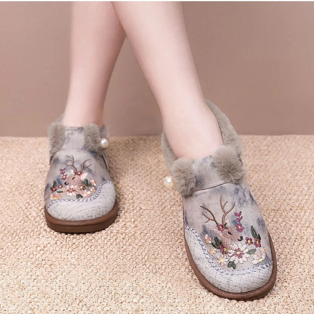 Embroidered plush slippers with grey faux fur trim and a pearl accent, worn by a person showing the floral and deer motif on a textured carpet background.