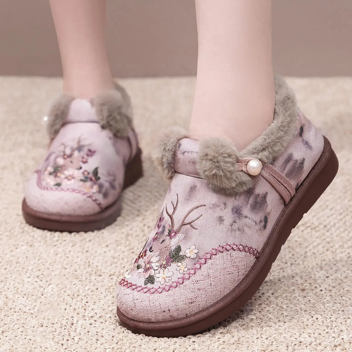 Embroidered plush slippers in a soft pinkish-mauve fabric with intricate floral and deer embroidery, featuring a grey faux fur trim and a single pearl accent.